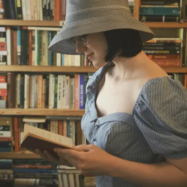 Vivian DeRosa with a gray hat on reads a book surrounded by bookshelves