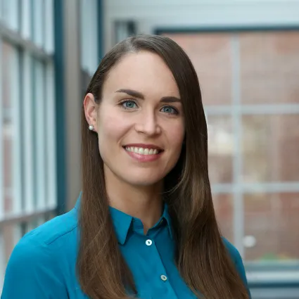 Headshot of a smiling woman with long brown hair parted on the side and light blue eyes. She is wearing a bright teal button-up shirt and pearl stud earrings, standing indoors in front of a soft-focused background with large window frames and brick detail.