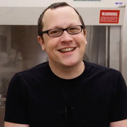 Portrait of a smiling man with short dark hair, black rectangular glasses, and a black t-shirt, standing in front of laboratory equipment with a red warning sign.