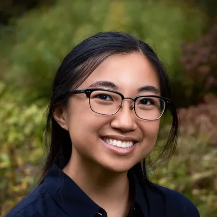 A headshot portrait of Juliet Ramey-Lariviere smiling at the camera while wearing dark-rimmed glasses and a dark blue collared shirt. The photo is set against a soft, out-of-focus background of green foliage.