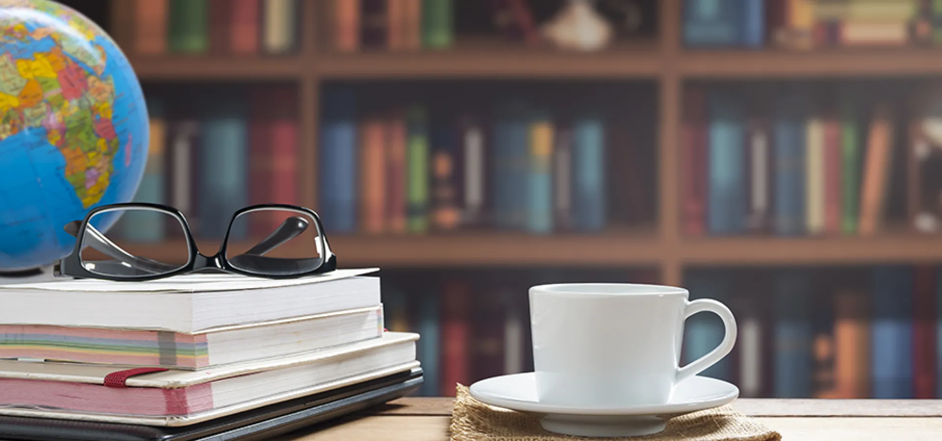 A globe centered on Africa sits on the corner of a desk. A pile of books is topped by a pair of glasses and to one side a saucer with coffee or tea. A vast bookcase covers the wall.