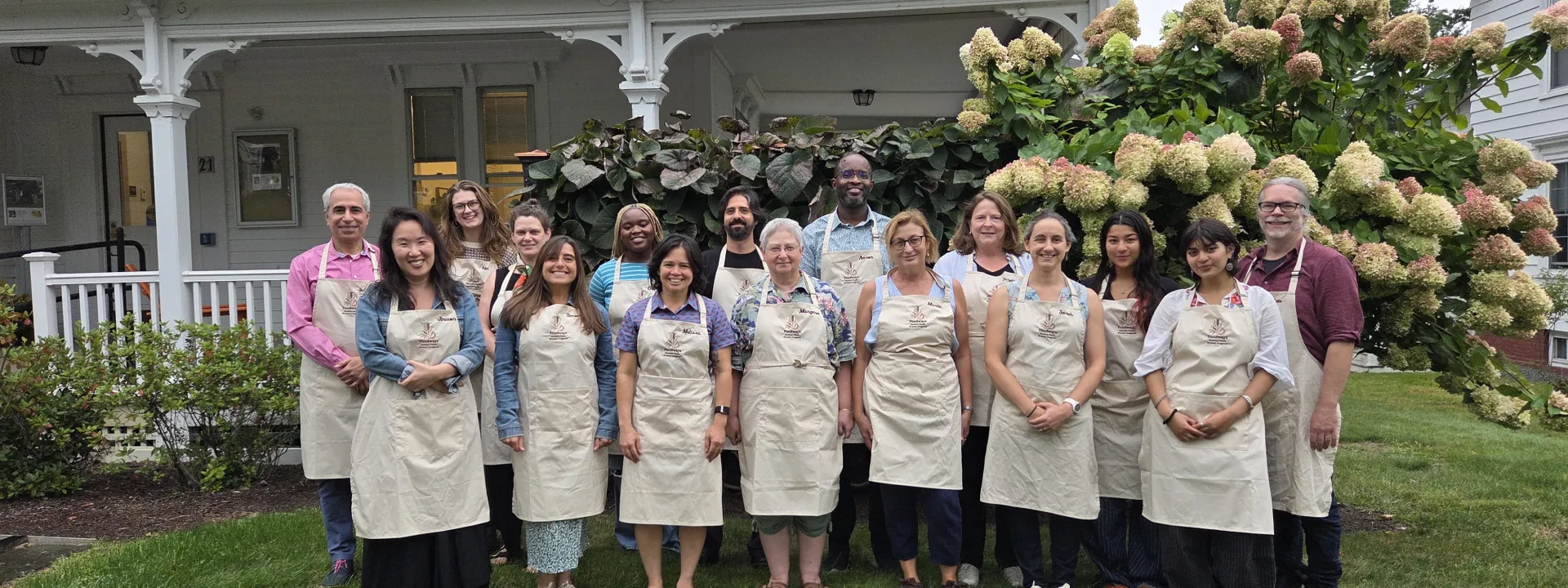 Group photo of fifteen Foodways fellows wearing matching cream aprons on a lawn outside a white house.