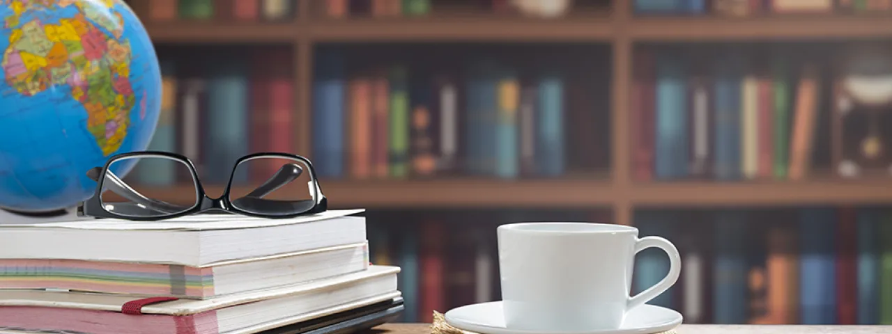 A globe centered on Africa sits on the corner of a desk. A pile of books is topped by a pair of glasses and to one side a saucer with coffee or tea. A vast bookcase covers the wall.