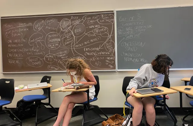 Two students writing, with a filled chalkboard behind them.