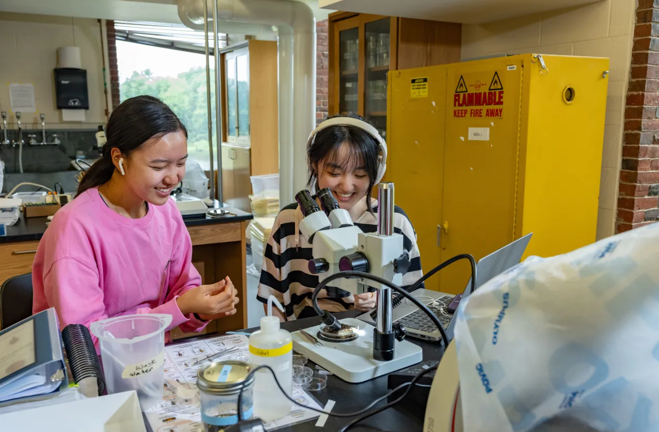 Two students, one in pink and one in stripes with headphones and earbuds, work at microscopes at Professor Marney Pratt's lab in Sabin-Reed