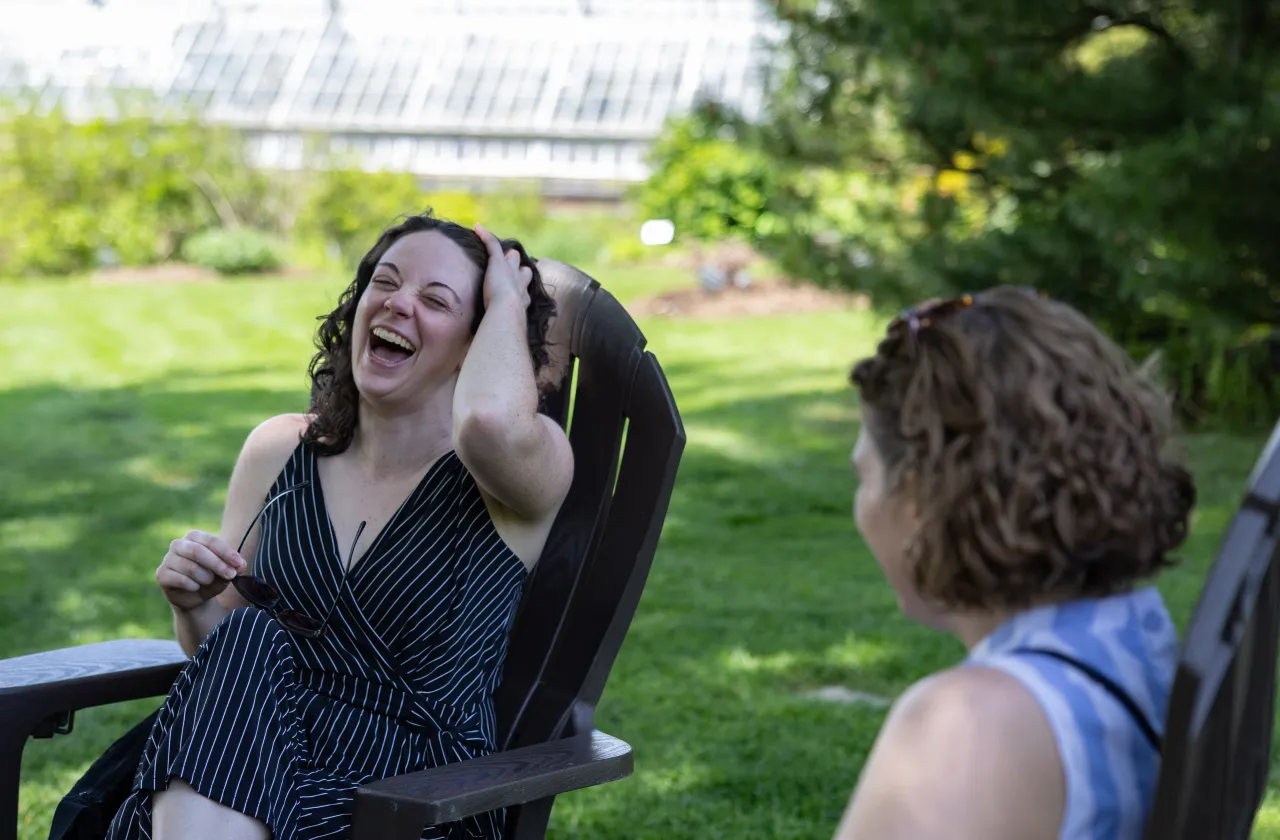 Two alums laugh in Adirondack chairs in front of the Lyman conservatory