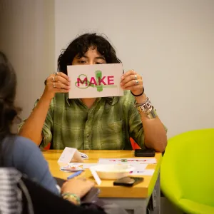 A student holds a paper with the words Make in front of their face while looking at the camera at ZineFest 2024