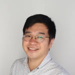 A portrait of Dong Woo Hahm smiling, with short black hair and glasses, and wearing a light checkered shirt.