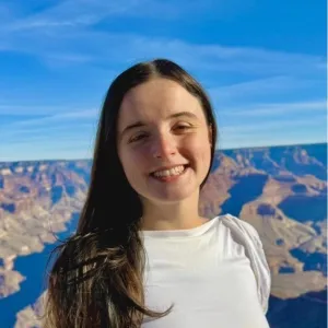 A smiling person with long dark hair stands in front of the Grand Canyon under a clear blue sky, wearing a white shirt. The scene conveys joy and serenity.