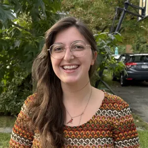 Outdoor headshot of Hannah smiling warmly, wearing round wire-frame glasses and a colorful patterned crochet sweater.