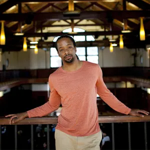 Jericho Brown leans on a wooden beam, looking directly into the camera. Photo credit: Stephanie Mitchell