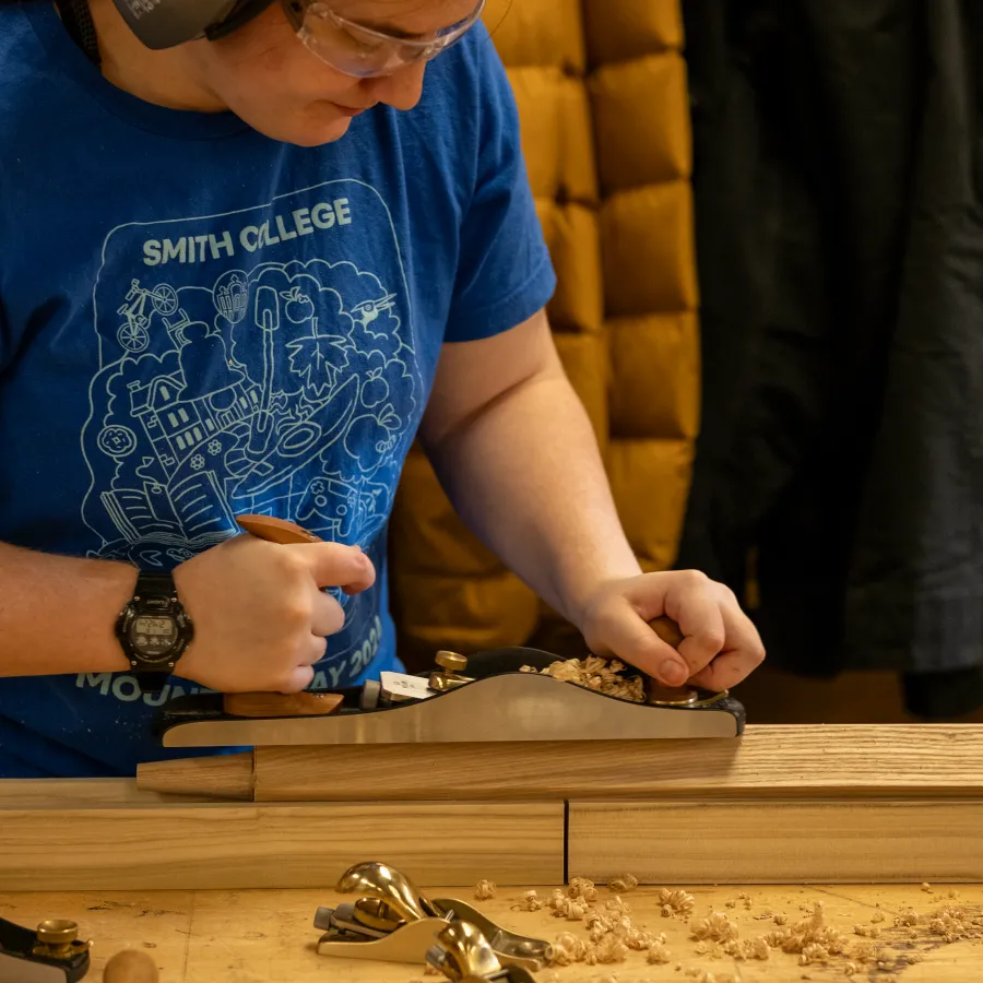 Elizabeth Rennert ‘25 in a blue 2024 Mountain Day shirt, pushes a Lie Neilson handplane across a block of wood