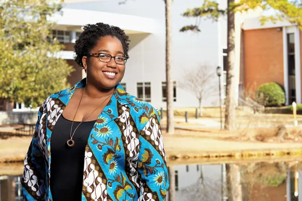 A black woman in a bright blue patterned jacket standing in front of a pond and brick building