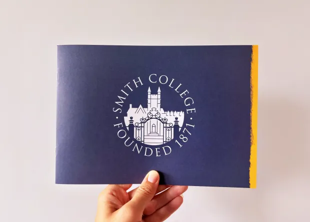 A hand holding a navy blue Smith College booklet featuring a white central crest and a gold edge against a neutral background.