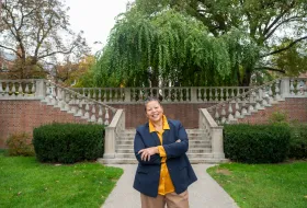 Photo of Sarah Willie-LeBreton smiling and standing outside with her arms folded, in front of the stone stairs in The Quad. 