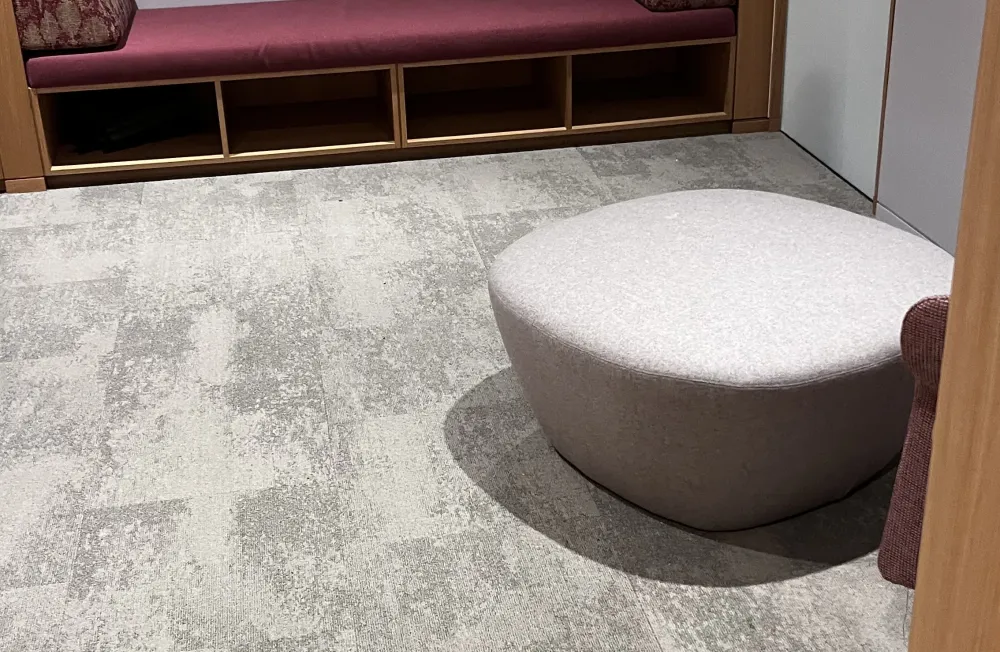 A cozy room features a purple cushioned bench with patterned side cushions against a wall, carpeted floor, and a round gray ottoman, creating a calm ambiance.