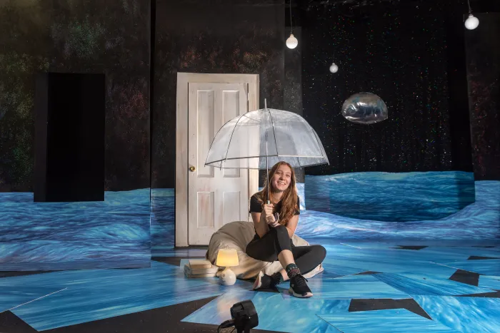 A student sitting on a stage holding an umbrella over her head.