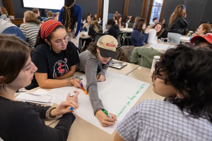 A group of students working together around a table. A student writes on a large white piece of paper with a green marker.