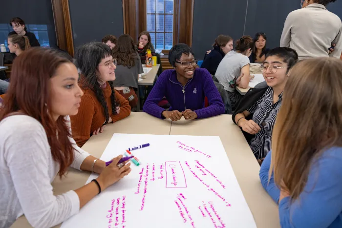 A group of students sits around a table, engaged in discussion, working together on a large white sheet of paper and writing with a purple marker