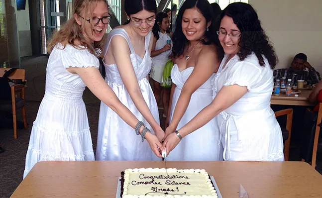 Four computer science grads in Commencement dresses cutting a cake