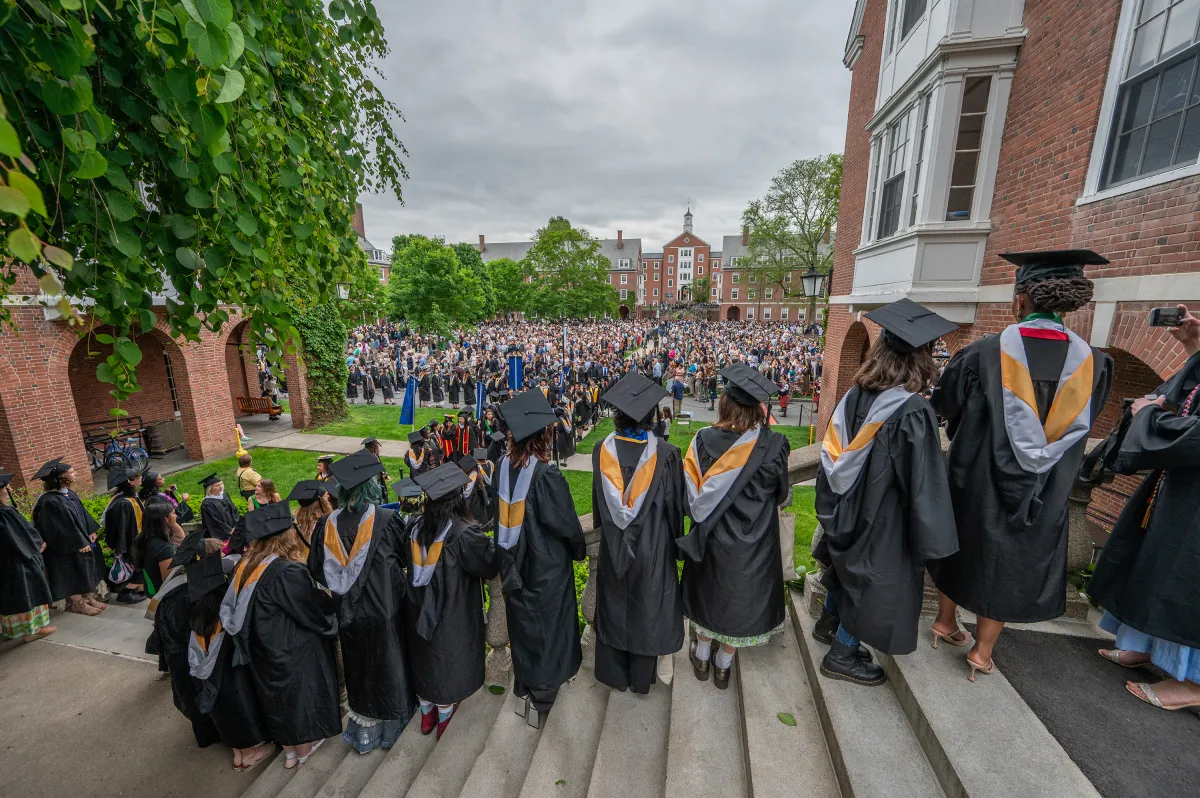 A group of seniors in caps and gowns with their backs facing the camera look out into the Quad on Commencement Day