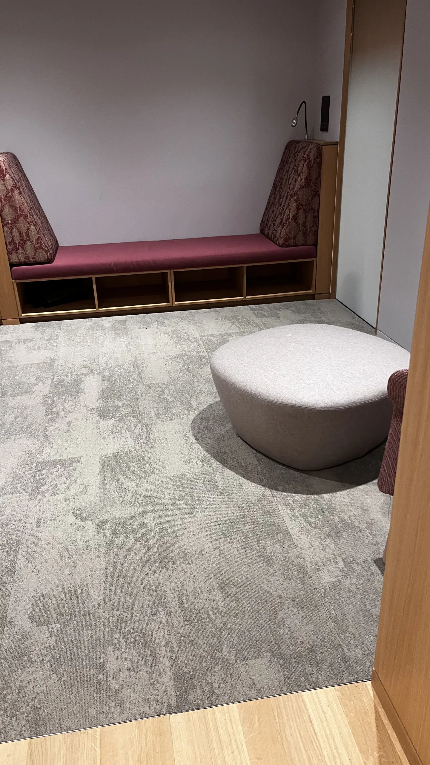 A cozy room features a purple cushioned bench with patterned side cushions against a wall, carpeted floor, and a round gray ottoman, creating a calm ambiance.
