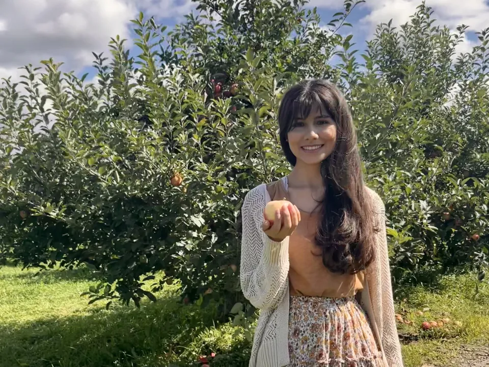 Gloria Parrales ’24, smiling, holds an apple in an orchard