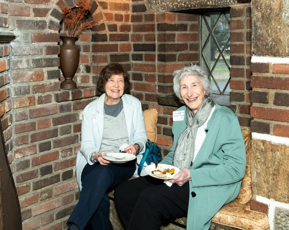 Two alums sit with plates of food talking with one another next to a rustic fireplace.