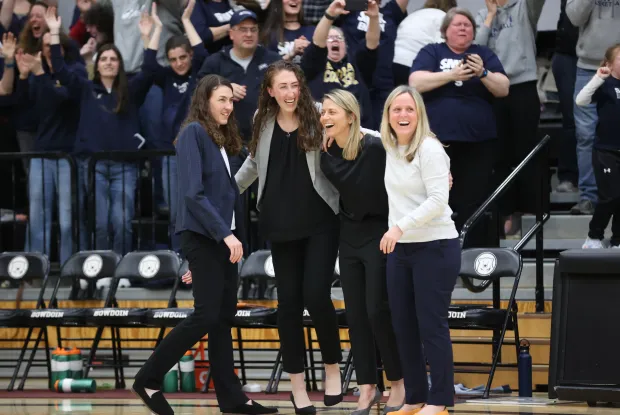 Four women on a basketball coaching staff celebrate joyfully in front of cheering fans during a game.