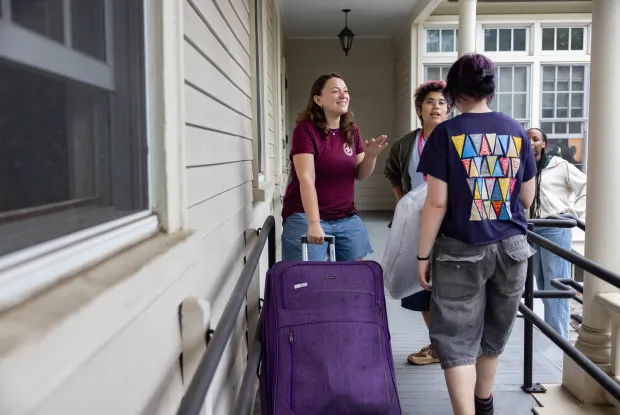 A student wheels a purple suitcase up a ramp to get into a house, while speaking animatedly to two other students